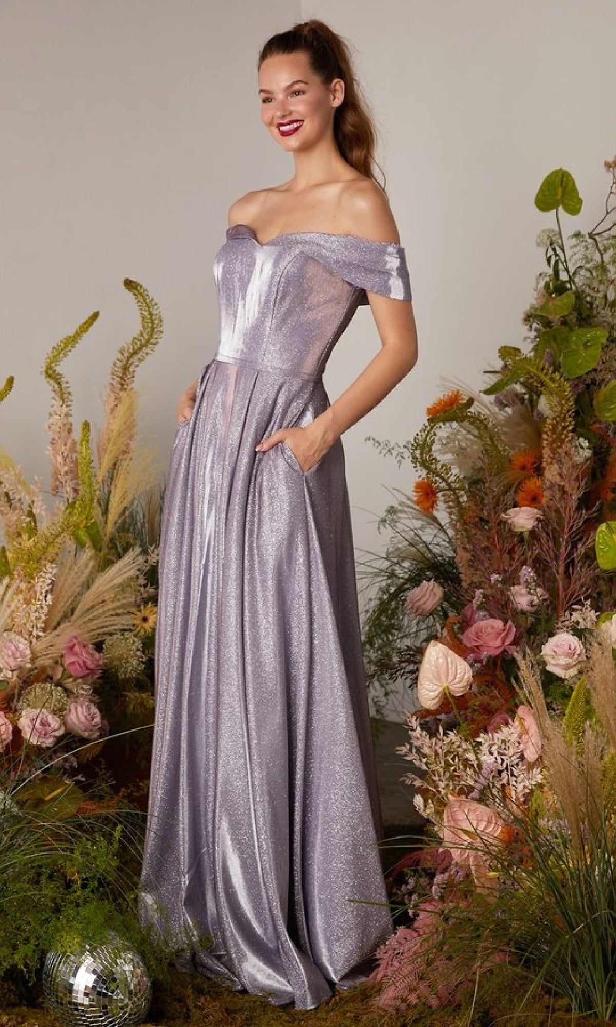 A woman stands among floral arrangements, smiling and looking to the side, wearing the shimmery lavender Eureka 9809 Off-Shoulder Long Glitter Prom Dress with pockets.